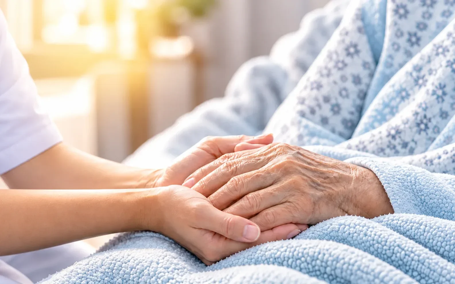 Hand hält die Hand einer schwer kranken Person unter einer hellblauen Decke – palliative Pflege